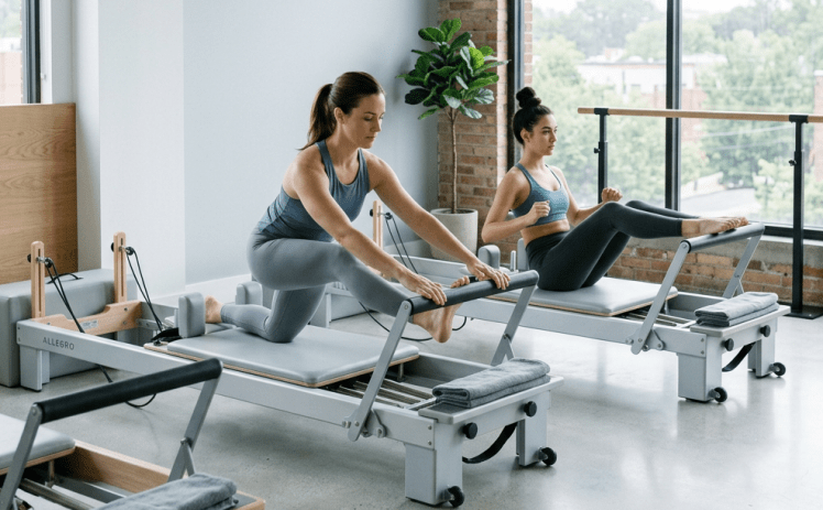 Two women doing Pilates exercises on reformer machines in a bright room with large windows